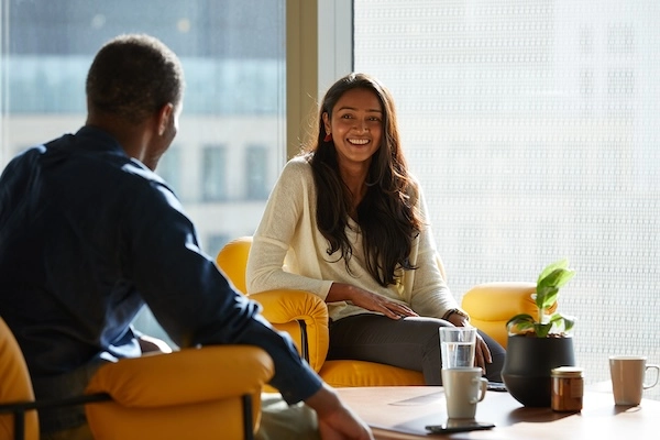 Two co-workers sit together around a coffee table enjoying a conversation and hot drink.
